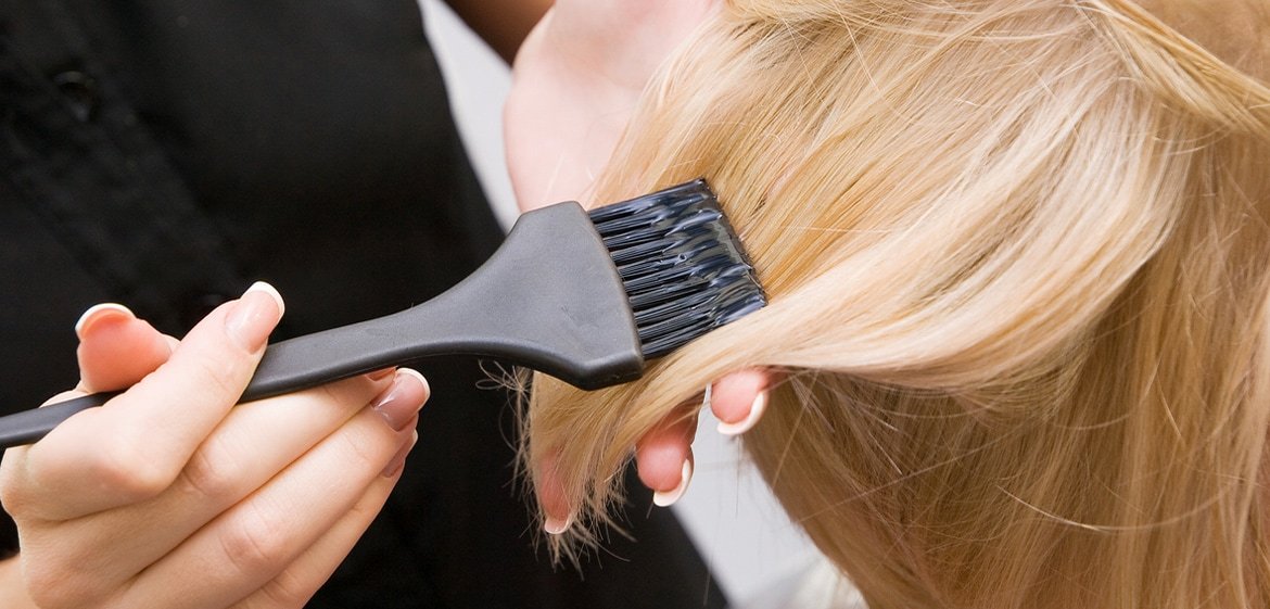 Ein kurzer, hellblonder Haarschöpf von schräg oben. Eine Frau trägt mit beiden Händen mittels eines schwarzen Pinsels Blondierung auf die Haare auf.
